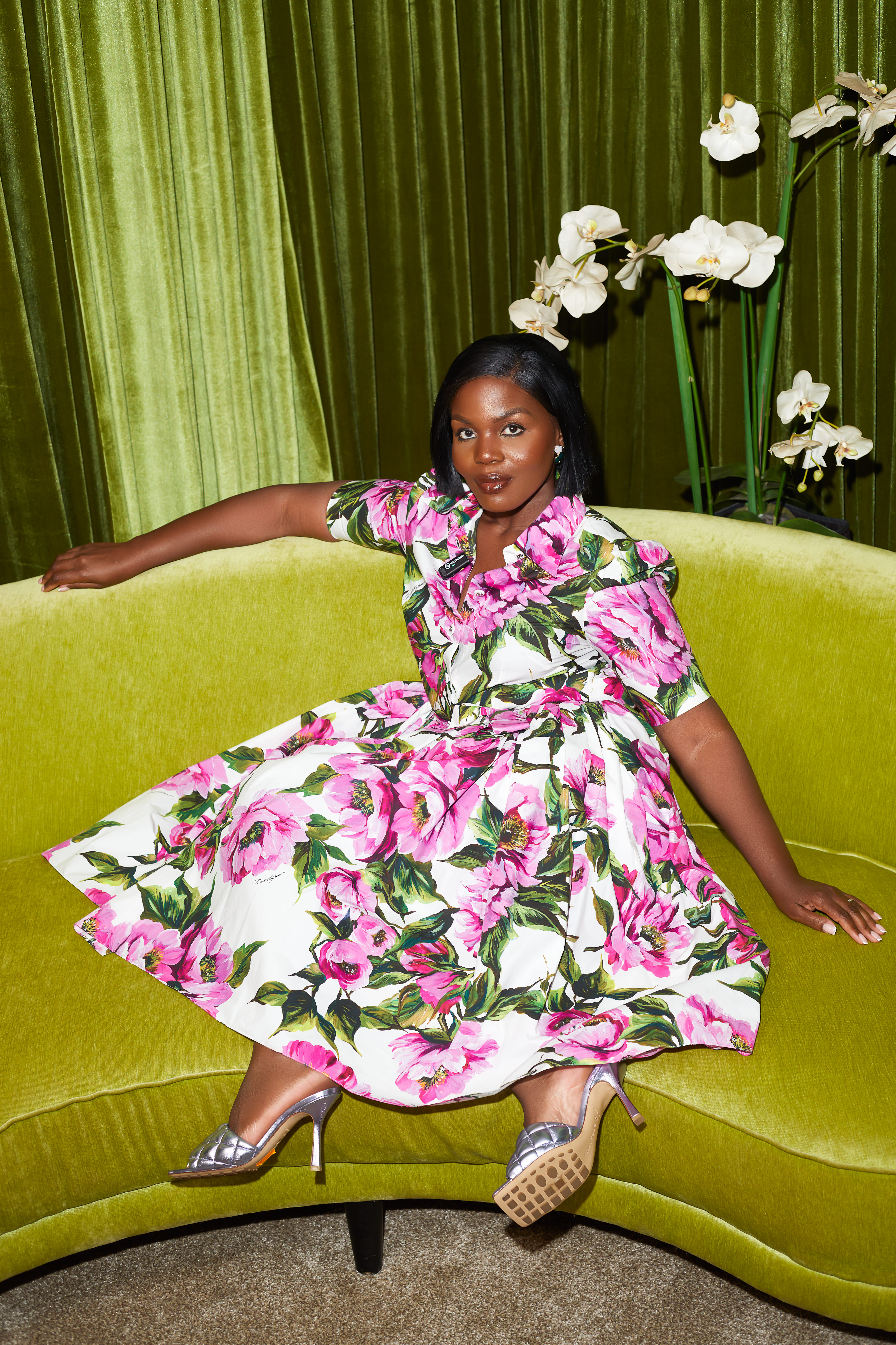 Sacha Okoh in a floral dress, seated on a green velvet sofa